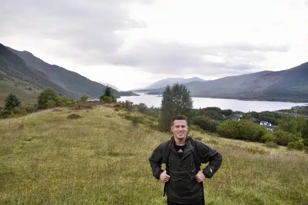 Kristoffer Fons di Destination Well Known su un sentiero erboso con un lago e le montagne sullo sfondo